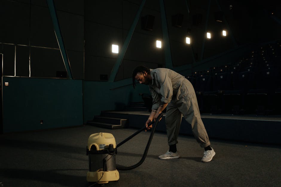 A person dressed in grey work overalls operating a yellow vacuum cleaner on a dark, carpeted floor in an auditorium or theatre setting. The room features tiered seating with numbered blue chairs, and the background includes dark walls with angular paneling and overhead lighting. The vacuum cleaner is emitting a mild light, indicating it is turned on, and the individual appears to be performing surface cleaning or deep cleaning to maintain hygiene and cleanliness in the space, demonstrating professional cleaning practices as offered by Brompton Carpet Cleaning.