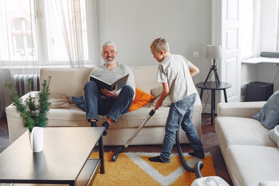 A young boy is vacuuming a plush orange and beige patterned rug in a well-lit living room, while an older man reclines on a cream-colored sofa nearby, appearing relaxed. The room features large window