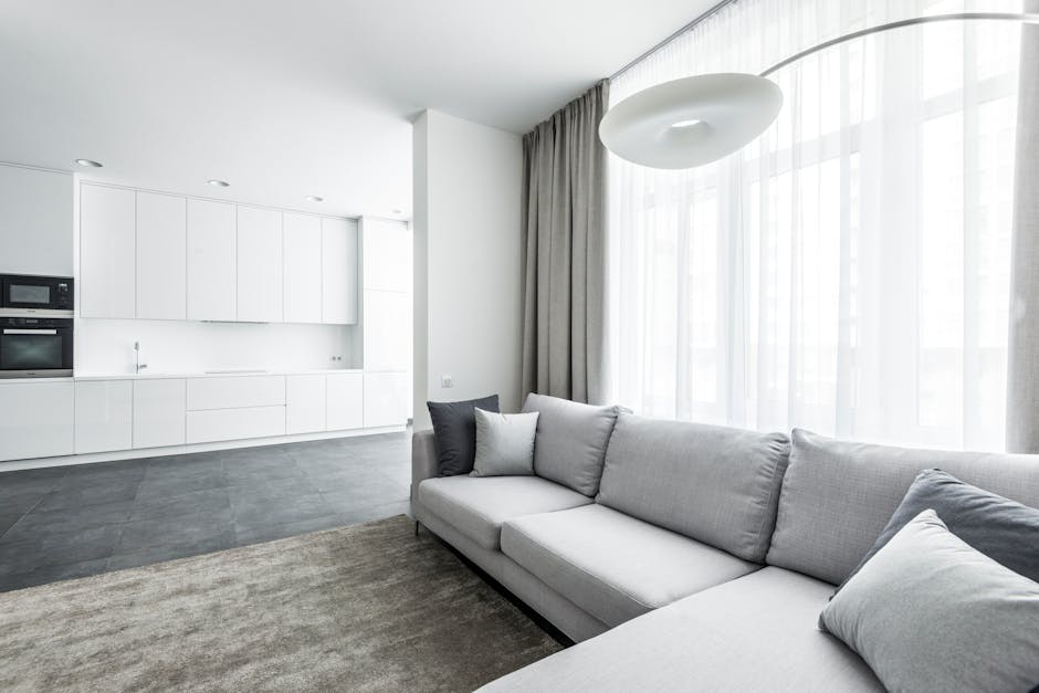 A modern, minimalist living room section with a light gray sofa adorned with dark and light gray cushions, positioned on a beige carpet. Behind the sofa, large windows with sheer white curtains allow natural light to fill the space. Adjacent to the living area, an open-concept kitchen features white cabinetry, integrated stainless steel appliances, and a dark gray tiled floor. The room is illuminated by natural daylight and a contemporary white ceiling light fixture. The overall appearance is clean, bright, and well-maintained, showcasing a neat and hygienic living space. Brompton Carpet Cleaning specializes in surface cleaning and deep cleaning services, ensuring enhanced hygiene and freshness in residential interiors like this.


